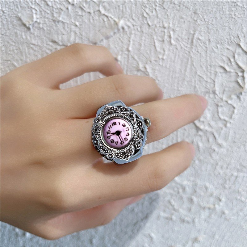 Ring-style watch with a pink dial on a person's finger, ornate silver band and decorative details against a textured wall background