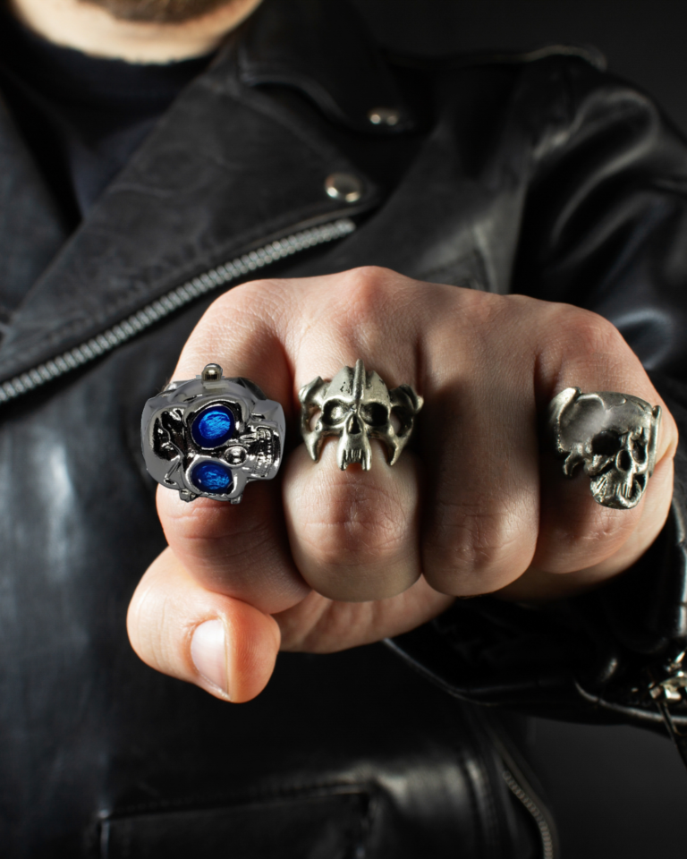 Skull-themed rings on a clenched hand, including a metallic skull ring with blue accents, against a black leather jacket background.